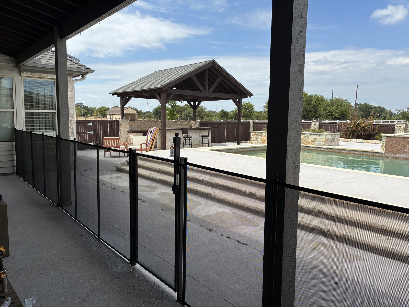 Black removable mesh pool fence with a self-closing gate off a covered patio, with the pool and pavilion beyond, in Georgetown TX